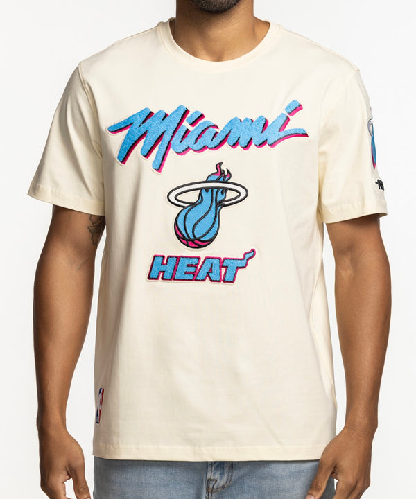 A man smiles with his hands in his pockets, wearing a Pro Standard Miami HEAT Original Vice Nights Logo Tee with neon pink and blue lettering, standing against a plain white background.
