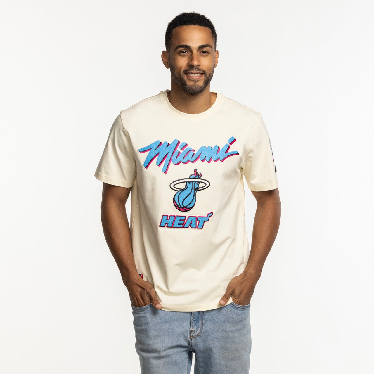 A man smiles with his hands in his pockets, wearing a Pro Standard Miami HEAT Original Vice Nights Logo Tee with neon pink and blue lettering, standing against a plain white background.