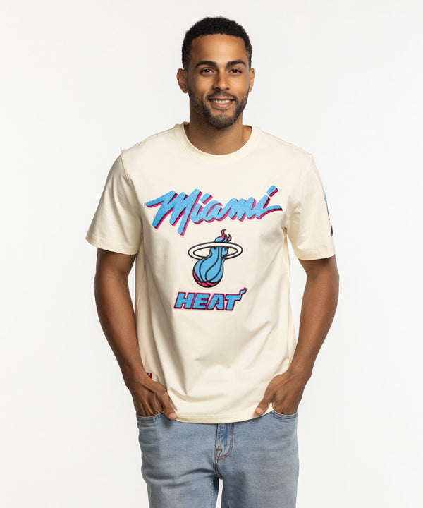 A man smiles with his hands in his pockets, wearing a Pro Standard Miami HEAT Original Vice Nights Logo Tee with neon pink and blue lettering, standing against a plain white background.