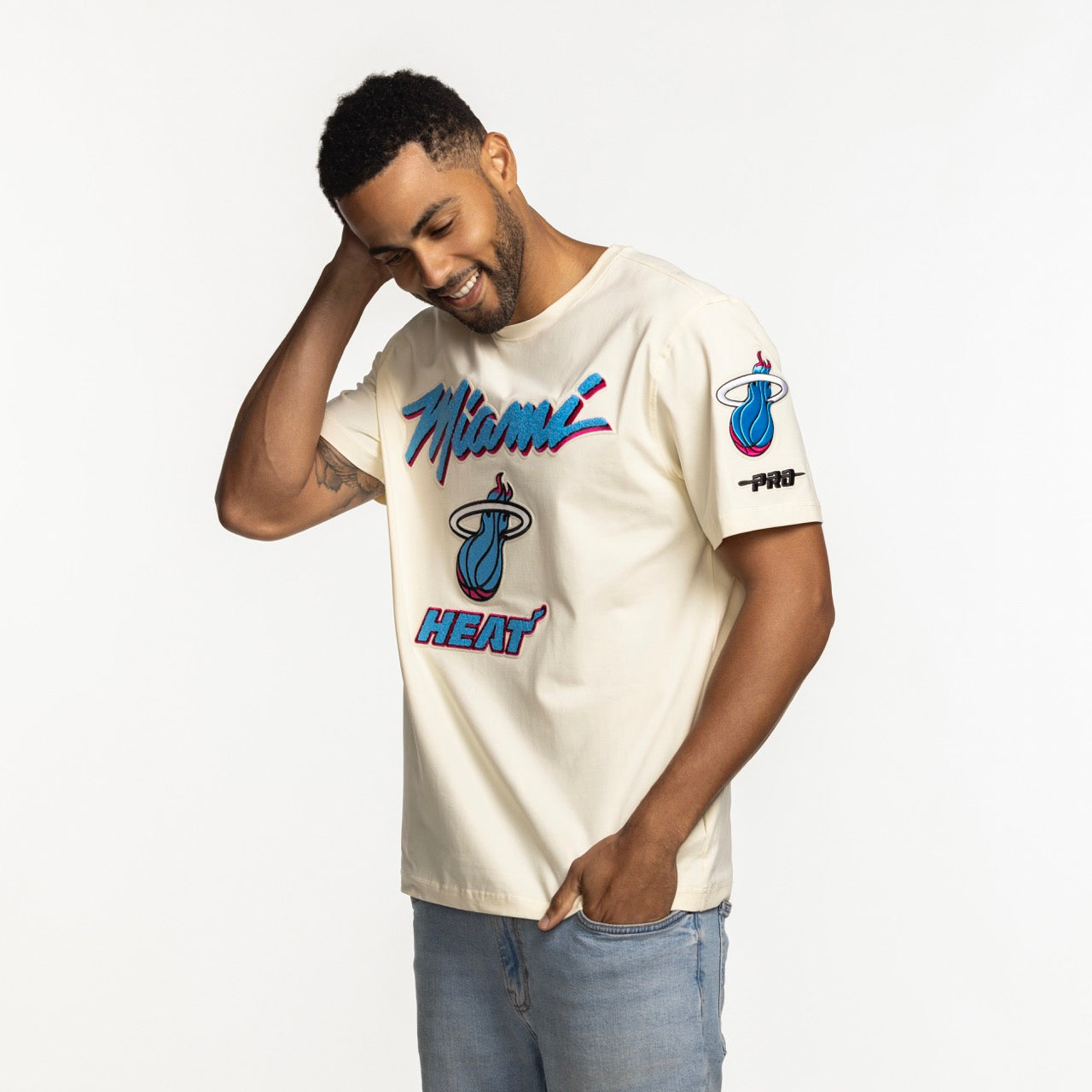 A man in a Pro Standard Miami HEAT Original Vice Nights Logo Tee, featuring team logos and blue/red accents in cream, standard fit, soft cotton spandex, stands smiling with one hand behind his head. Machine washable. Brand: Pro Standard.