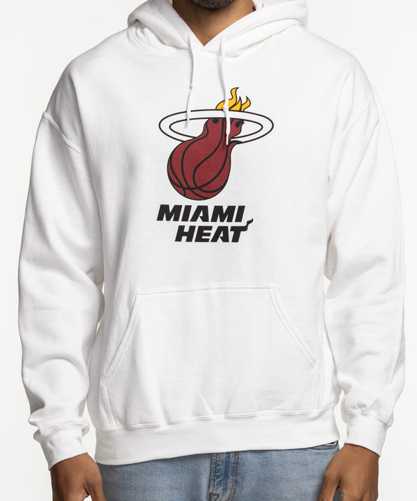 A man smiles with hands in pockets, wearing the Miami HEAT Logo White Hoodie by Item Of The Game and light blue jeans. The white cotton-poly hoodie features the team logo/text on front. Standard fit; machine wash cold for care.