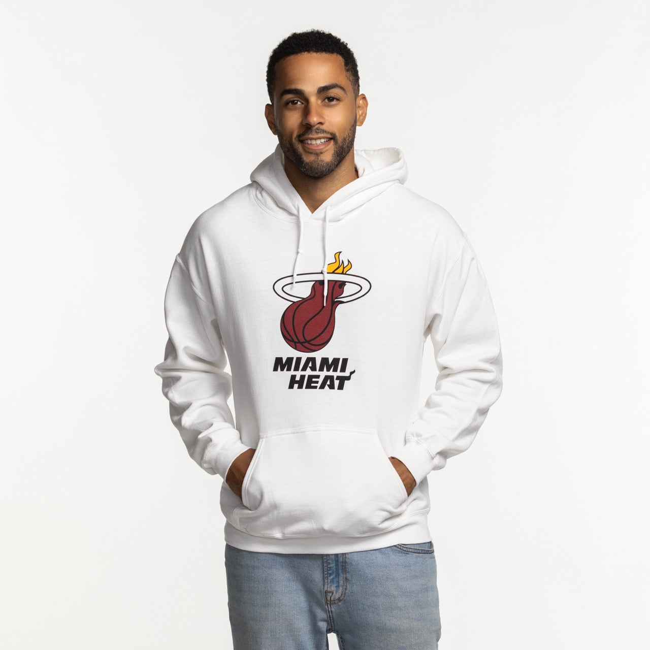 A man smiles with hands in pockets, wearing the Miami HEAT Logo White Hoodie by Item Of The Game and light blue jeans. The white cotton-poly hoodie features the team logo/text on front. Standard fit; machine wash cold for care.