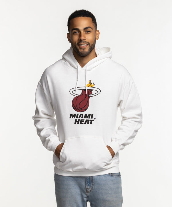 A man smiles with hands in pockets, wearing the Miami HEAT Logo White Hoodie by Item Of The Game and light blue jeans. The white cotton-poly hoodie features the team logo/text on front. Standard fit; machine wash cold for care.