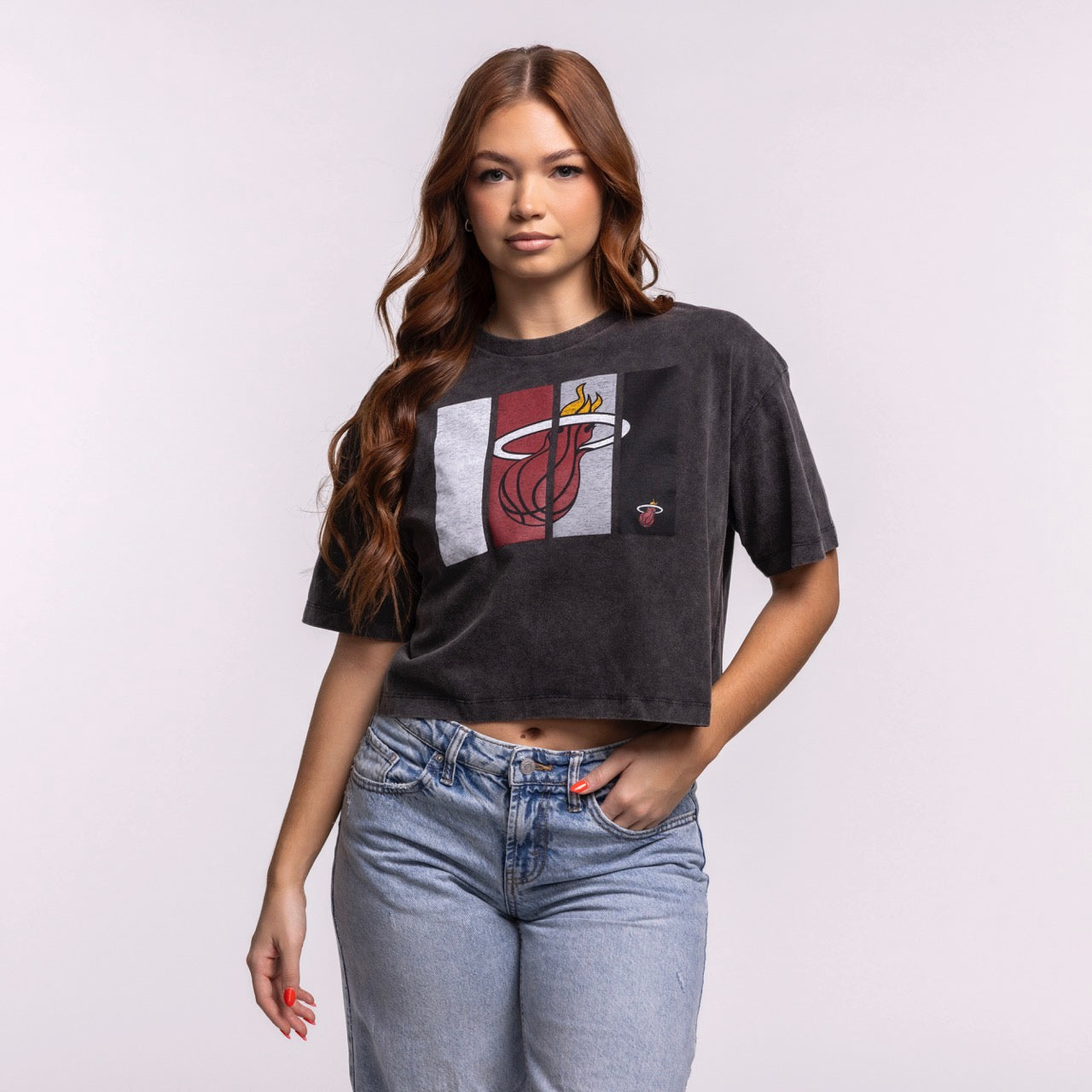 A woman with long brown hair wears the New Era Miami HEAT Washed Women's Cropped Tee—black and cotton—paired with light blue jeans, standing against a plain light background.