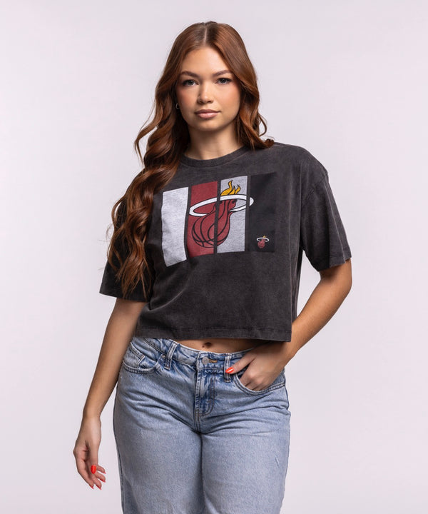 A woman with long brown hair wears the New Era Miami HEAT Washed Women's Cropped Tee—black and cotton—paired with light blue jeans, standing against a plain light background.