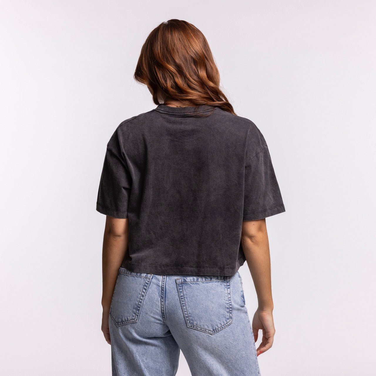 A woman with long, wavy brown hair is seen from the back, wearing the New Era Miami HEAT Washed Women's Cropped Tee in dark gray, paired with light blue high-waisted jeans, set against a plain light background.