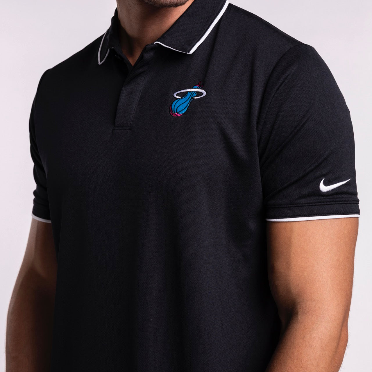 A person wears a black Nike Miami HEAT Original Vice Nights Logo Polo with white trim and team logo on the chest. The standard fit shirt is 100% polyester, shown in size L. The person's face is partially out of frame.