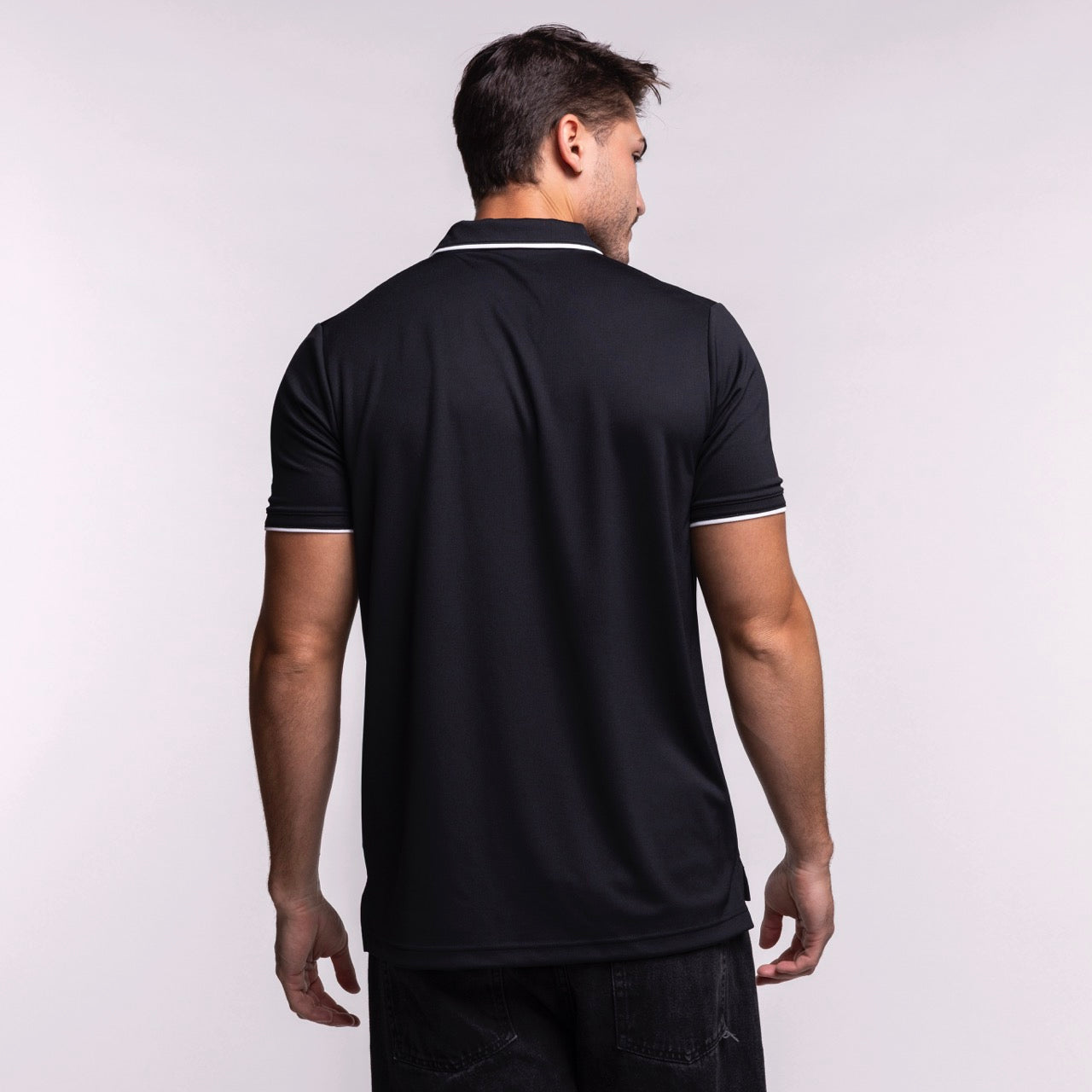 A man faces away from the camera, wearing a size L Nike Miami HEAT Original Vice Nights Logo Polo—a short-sleeved black shirt with white trim, 100% polyester, standard fit—paired with dark pants against a plain light background.