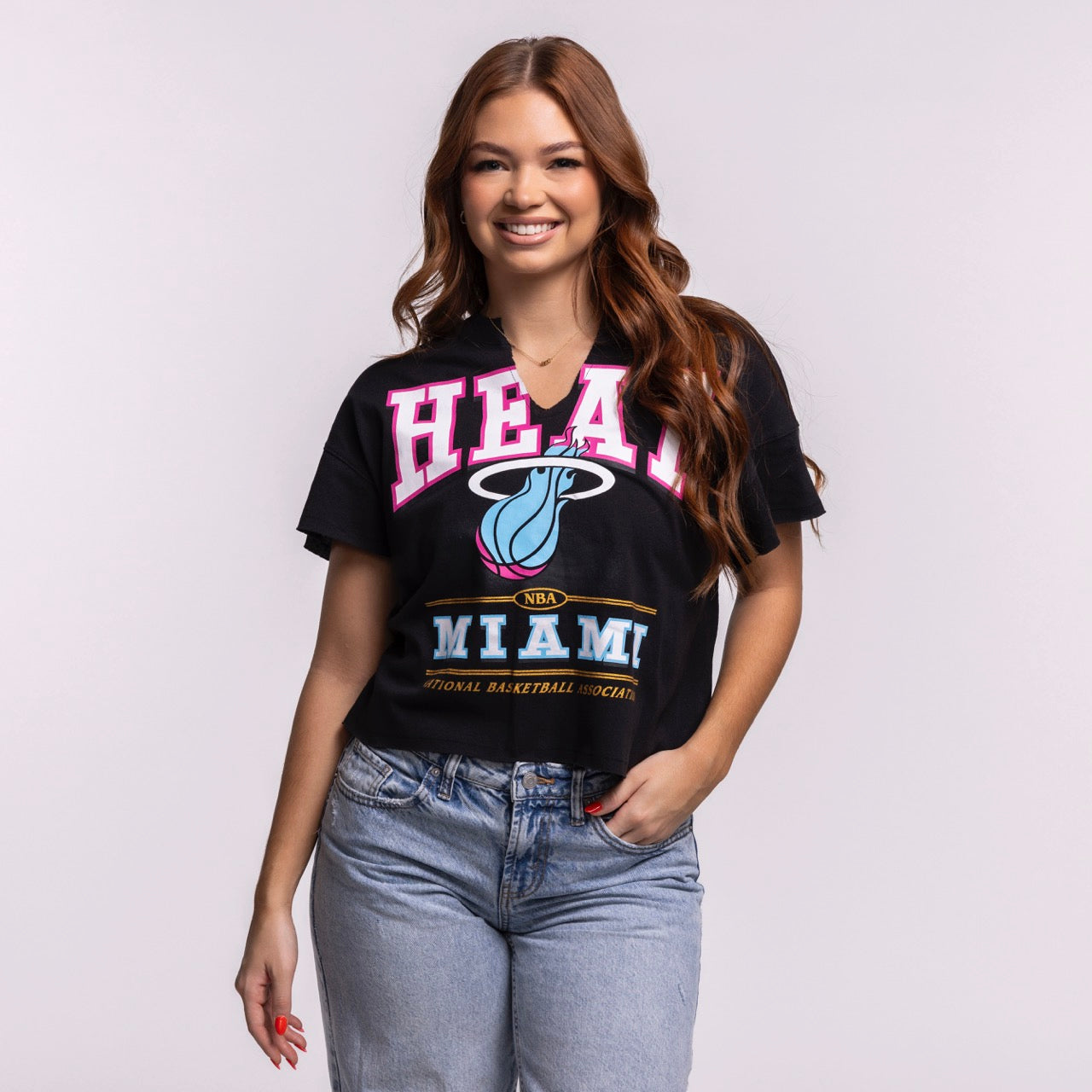 A woman with long, wavy brown hair smiles in a '47 Brand Miami HEAT Original Vice Nights Oversized Women's Cropped Tee and light blue jeans, standing against a plain background—a chic nod to women's fashion.