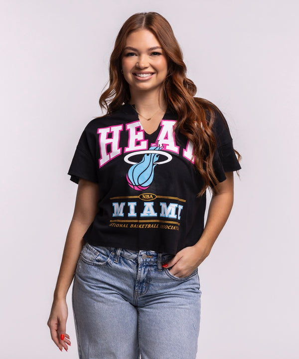 A woman with long, wavy brown hair smiles in a '47 Brand Miami HEAT Original Vice Nights Oversized Women's Cropped Tee and light blue jeans, standing against a plain background—a chic nod to women's fashion.