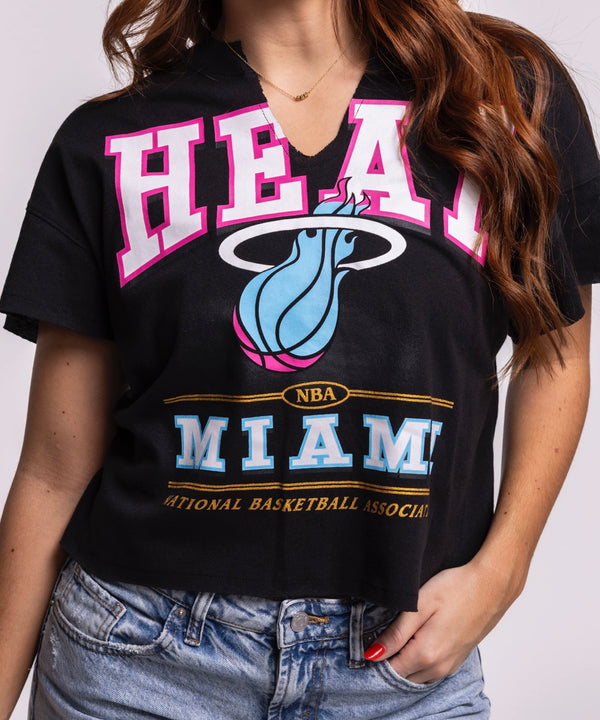 A woman with long, wavy brown hair smiles in a '47 Brand Miami HEAT Original Vice Nights Oversized Women's Cropped Tee and light blue jeans, standing against a plain background—a chic nod to women's fashion.
