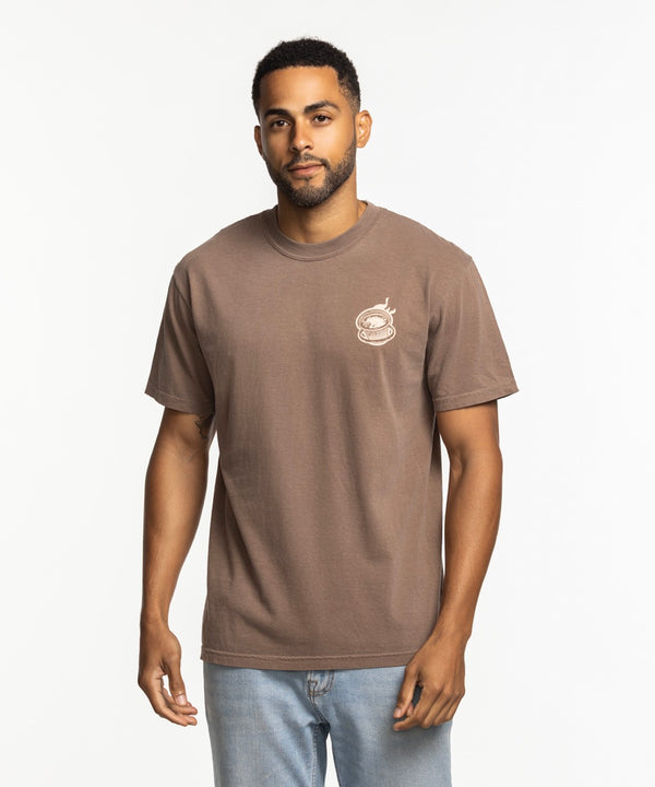 A man with short hair and a beard wears the Court Culture Burnie's Ventanita Espresso Unisex Tee by Court Culture, featuring a relaxed fit and small white chest graphic, with light blue jeans against a plain white background.