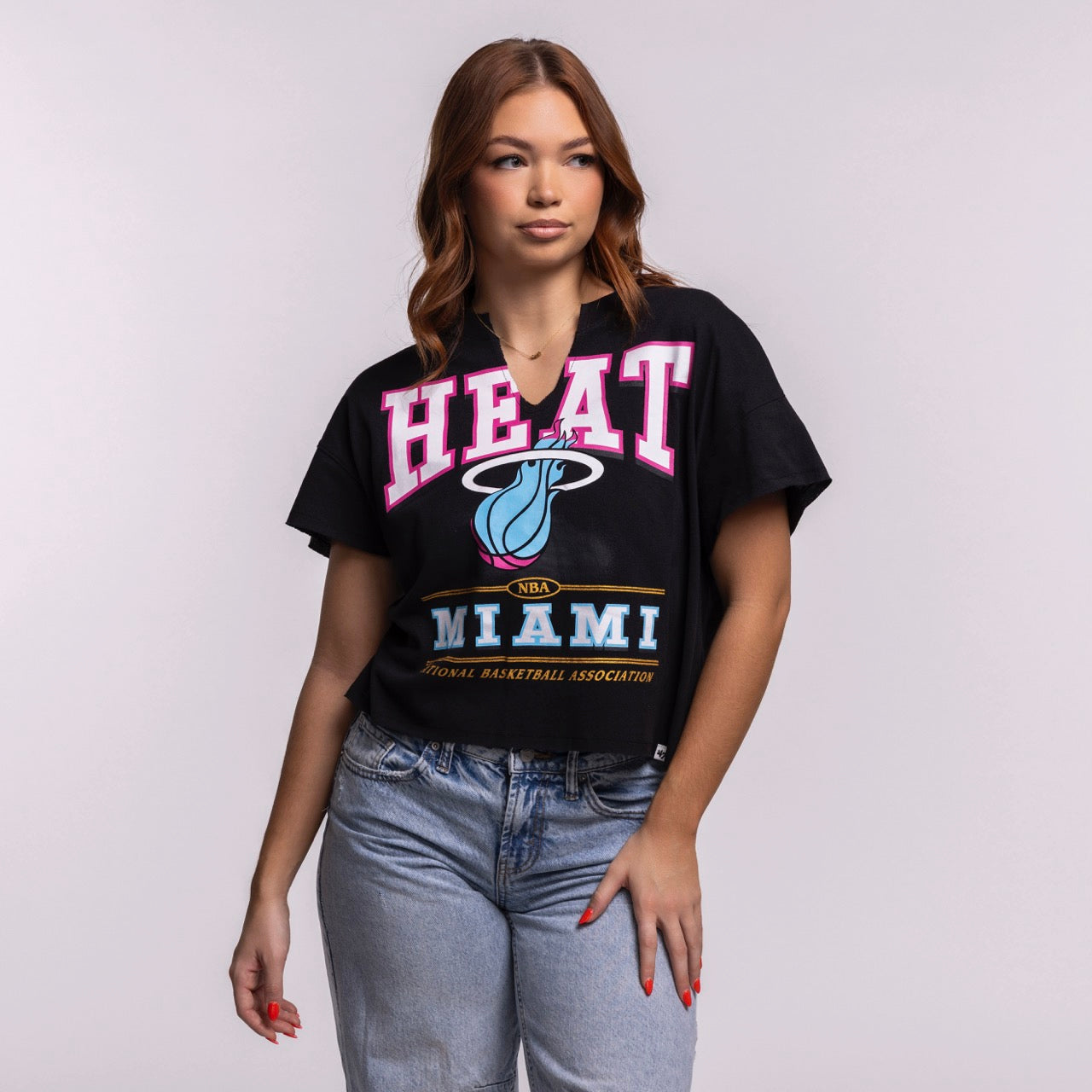 A woman with wavy brown hair wears the '47 Brand Miami HEAT Original Vice Nights Oversized Women's Cropped Tee and light blue jeans, standing against a plain background—a trendy choice for women's fashion.