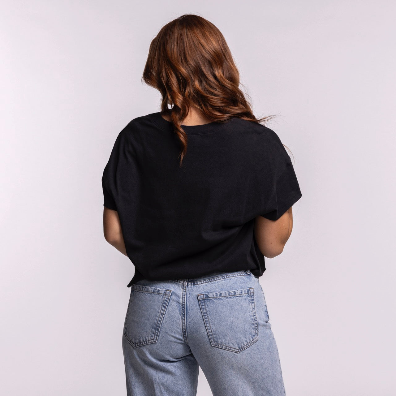 A woman with long wavy brown hair, wearing the '47 Brand Miami HEAT Original Vice Nights Oversized Women's Cropped Tee in black and light blue jeans, stands facing away from the camera against a plain light background—effortless style.