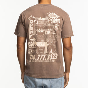 A person wearing the Court Culture Burnie's Ventanita Espresso Unisex Tee, a relaxed fit brown cotton shirt with a vintage Cuban cafe graphic, "Cafe Cubano," "Open" text, and Miami, FL address and phone number on the back.
