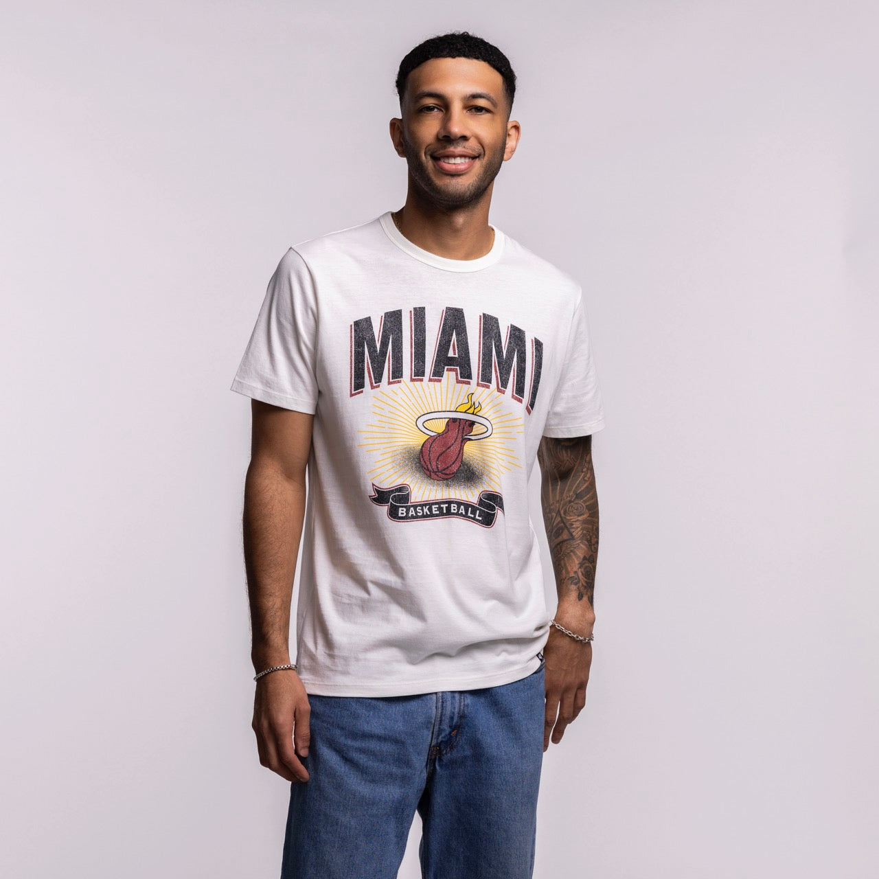A man smiles in front of a plain light background wearing the '47 Brand Miami HEAT Play Basketball Tee, a white, standard fit cotton shirt with a Miami basketball logo, paired with blue jeans. This '47 Brand tee is machine washable for easy care.