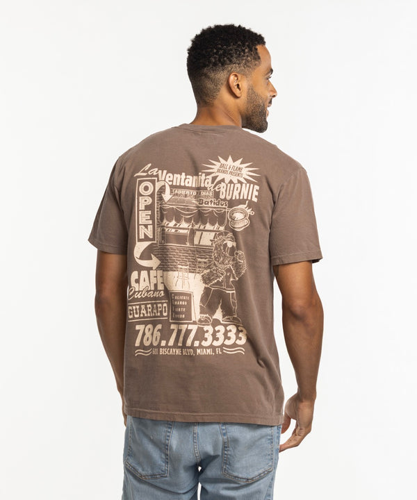 A man with short hair and a beard wears the Court Culture Burnie's Ventanita Espresso Unisex Tee by Court Culture, featuring a relaxed fit and small white chest graphic, with light blue jeans against a plain white background.