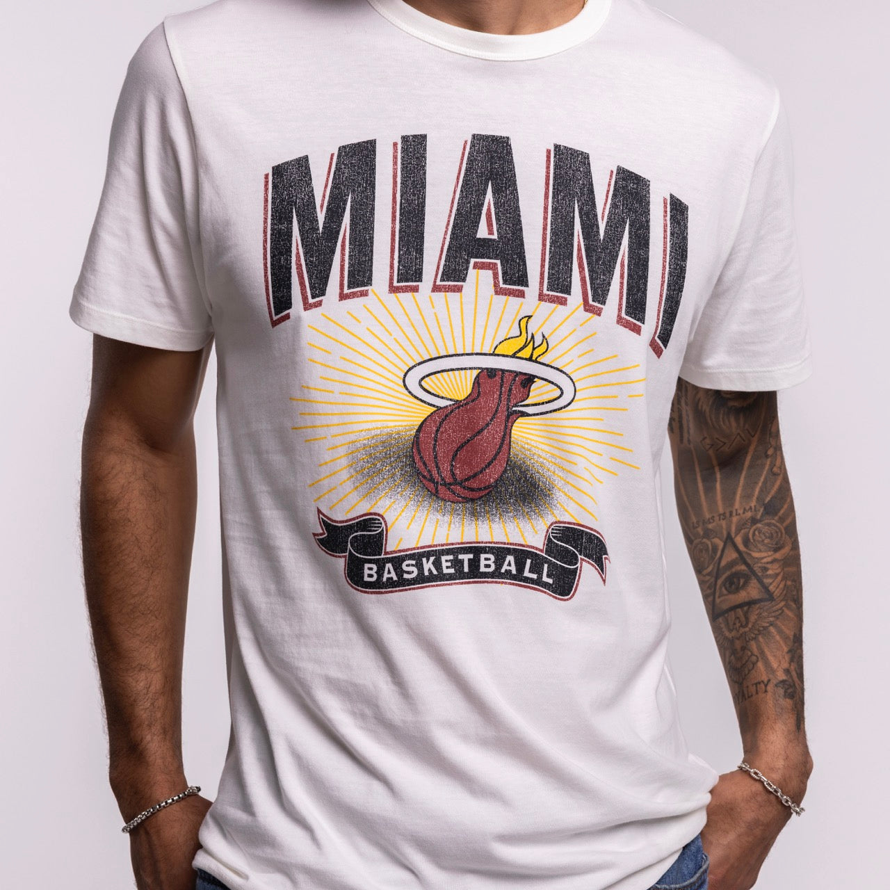 A person wearing a standard fit, white ‘47 Brand Miami HEAT Play Basketball Tee, featuring large MIAMI text, a flaming basketball logo, and BASKETBALL ribbon. Machine washable tee styled with tattoos on one arm and hands in pockets.