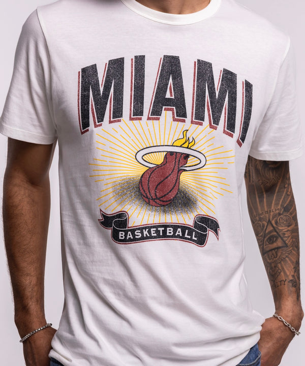 A man in a '47 Brand Miami HEAT Play Basketball Tee and blue jeans stands against a plain, light background, looking to his left.