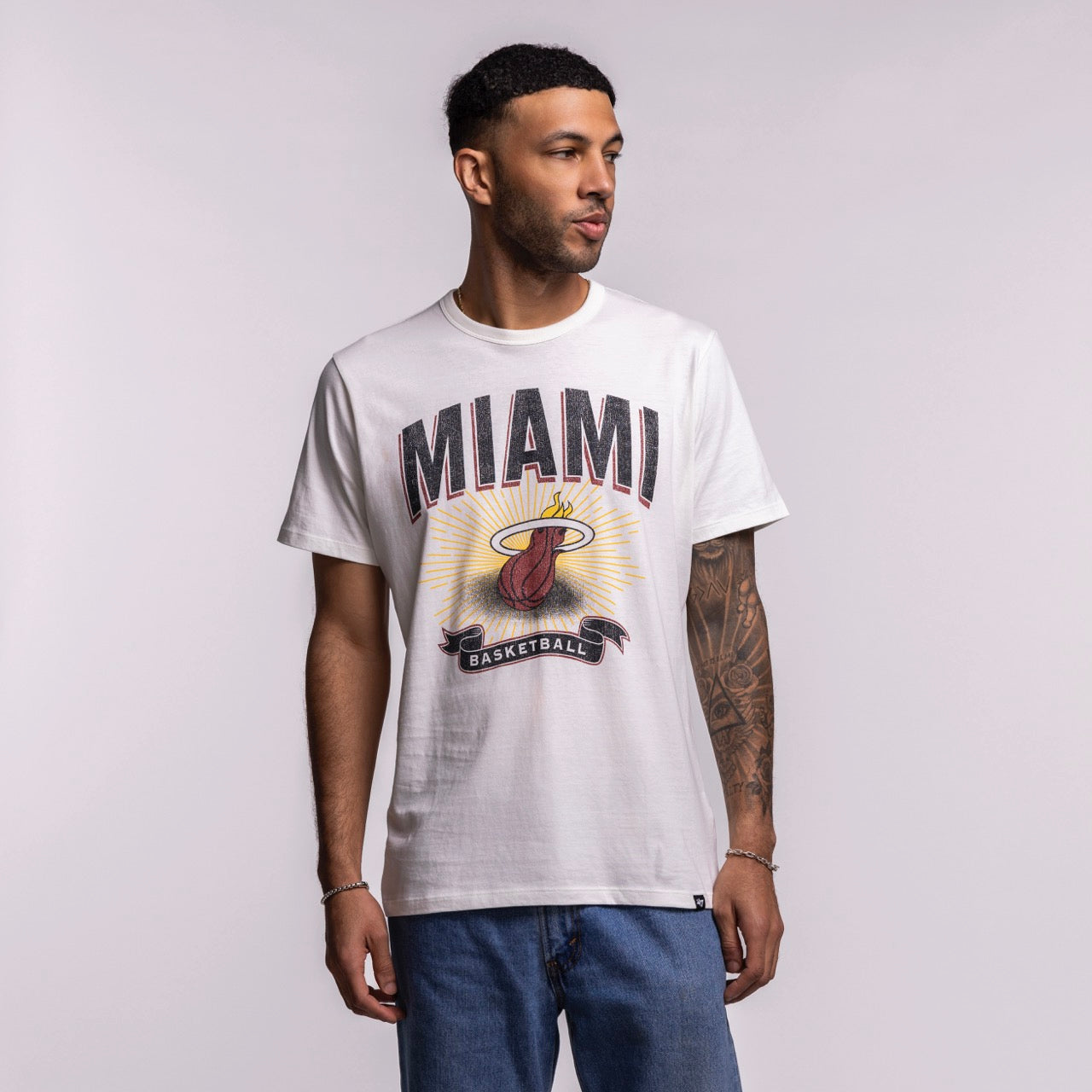 A man in a '47 Brand Miami HEAT Play Basketball Tee and blue jeans stands against a plain, light background, looking to his left.