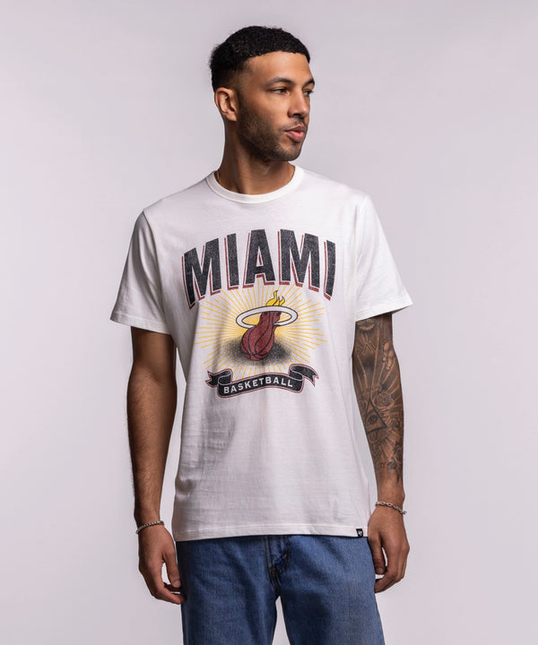 A man in a '47 Brand Miami HEAT Play Basketball Tee and blue jeans stands against a plain, light background, looking to his left.