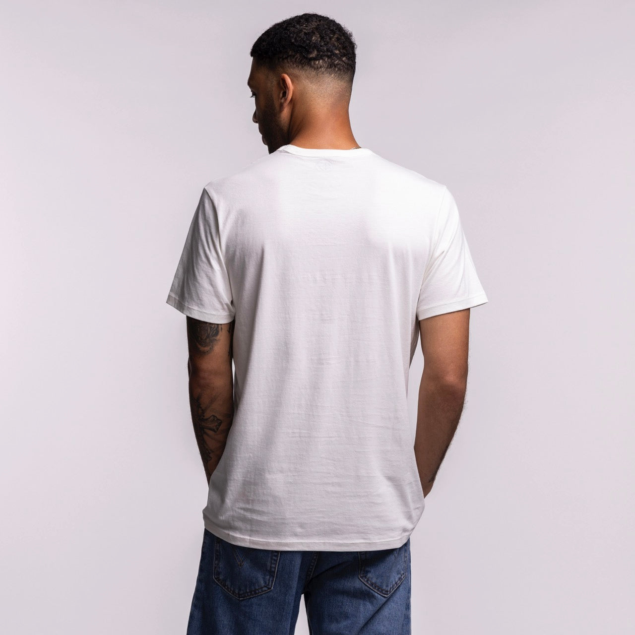 A man with short hair and arm tattoos is seen from the back, wearing a '47 Brand Miami HEAT Play Basketball Tee and blue jeans, set against a light gray background.