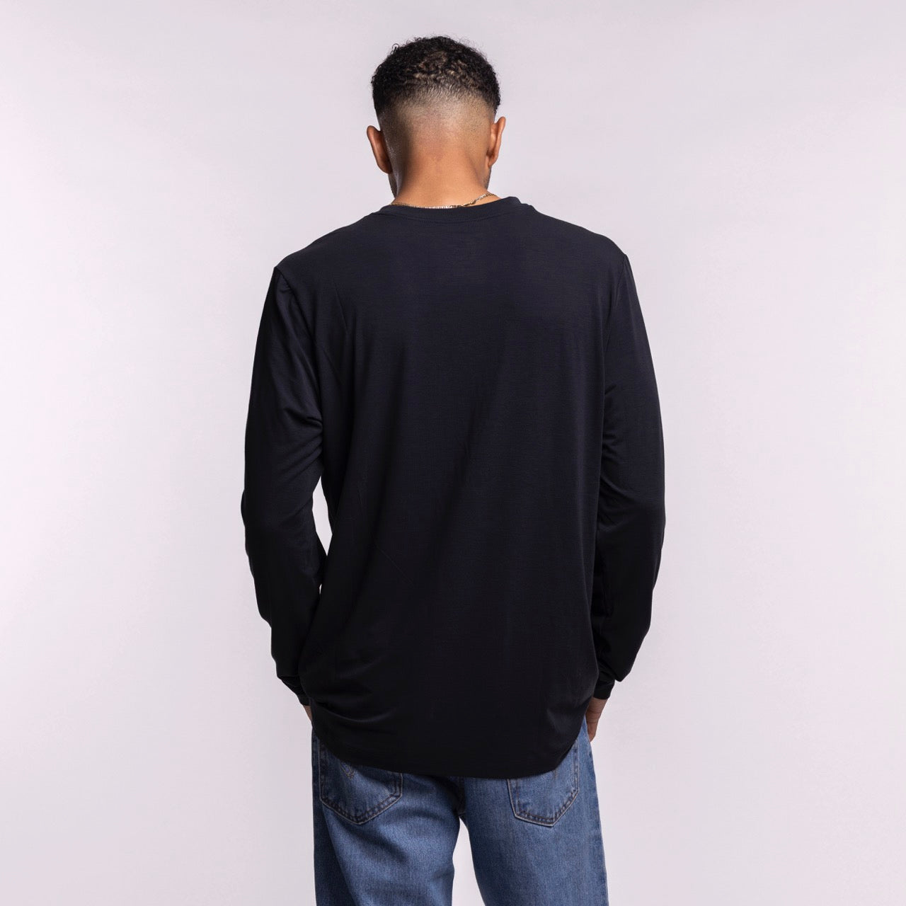 A man stands with hands in pockets, head slightly bowed, wearing a Nike Miami HEAT 2025-26 Authentic On-Court Practice Long Sleeve Tee and blue jeans against a plain light background, facing away from the camera.