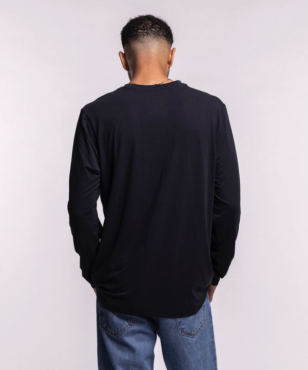 A man with short hair and a trimmed beard wears blue jeans and the Nike Miami HEAT 2025-26 Authentic On-Court Practice Long Sleeve Tee, featuring NBA and HEAT logos, against a light gray background.