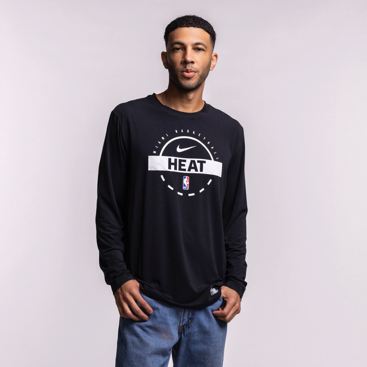 A man with short hair and a trimmed beard wears blue jeans and the Nike Miami HEAT 2025-26 Authentic On-Court Practice Long Sleeve Tee, featuring NBA and HEAT logos, against a light gray background.