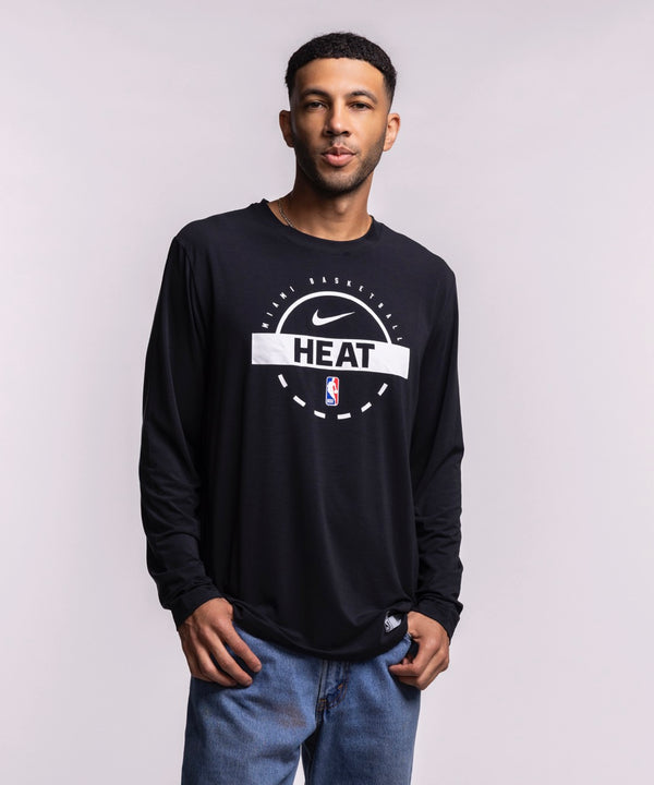 A man with short hair and a trimmed beard wears blue jeans and the Nike Miami HEAT 2025-26 Authentic On-Court Practice Long Sleeve Tee, featuring NBA and HEAT logos, against a light gray background.