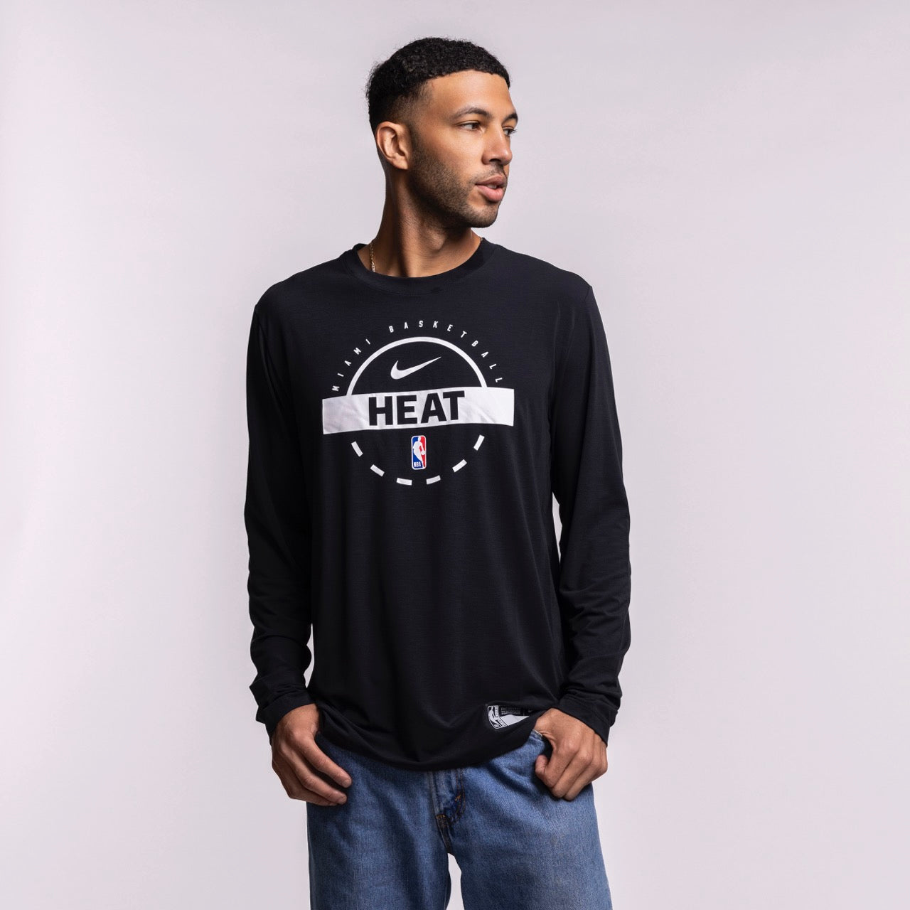 A man in a Nike Miami HEAT 2025-26 Authentic On-Court Practice Long Sleeve Tee made of soft polyester and blue jeans stands against a plain light background, looking to his left with his hands in his pockets.