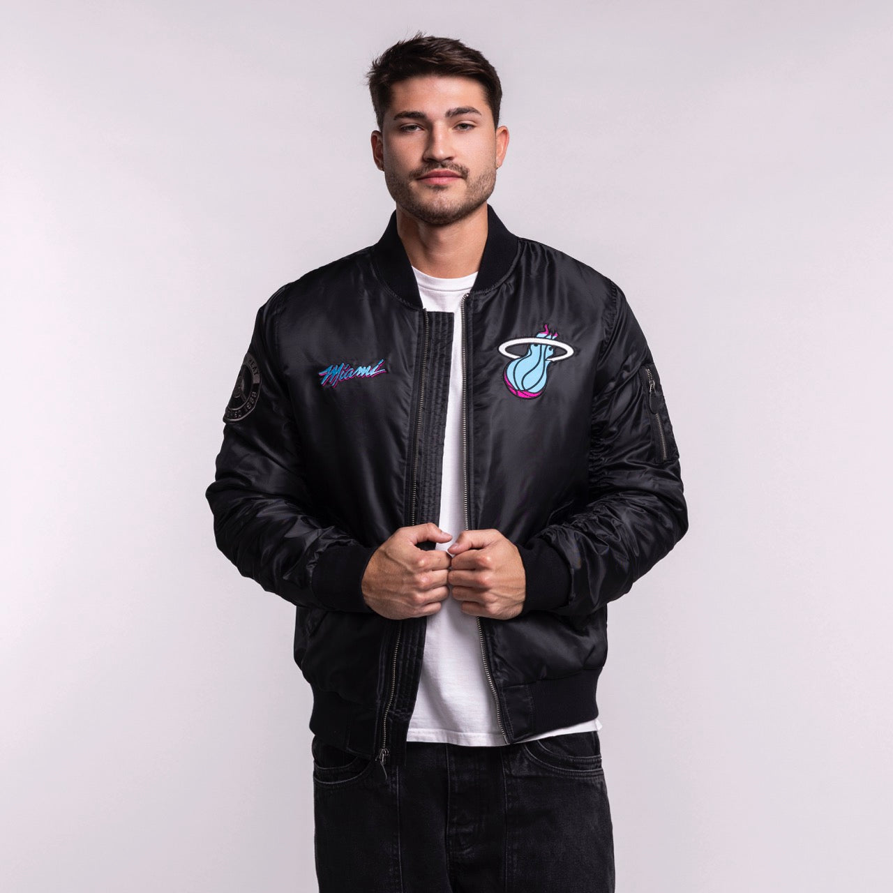 A man models the G-III Sports Miami HEAT Original Vice Nights Aviator Jacket in men's size L, standing against a plain light background and looking at the camera while holding the polyester, standard fit jacket front. Machine wash cold.