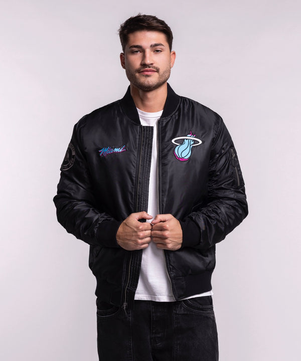 A man models the G-III Sports Miami HEAT Original Vice Nights Aviator Jacket in men's size L, standing against a plain light background and looking at the camera while holding the polyester, standard fit jacket front. Machine wash cold.