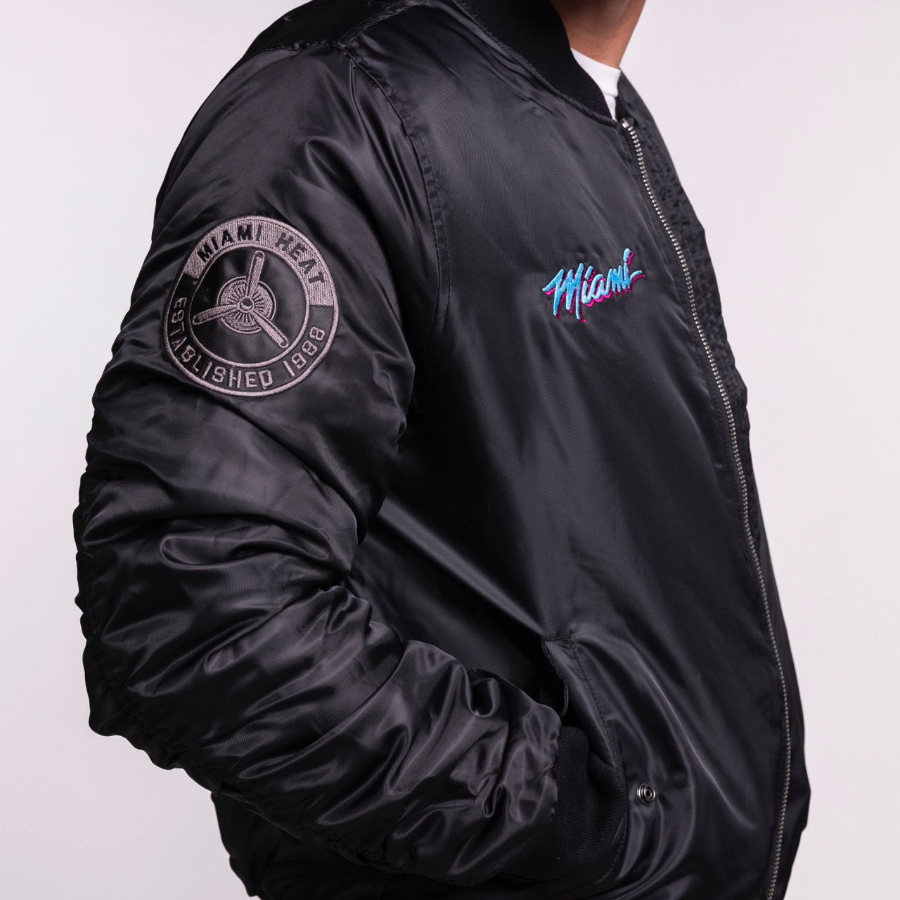 A person in a G-III Sports Miami HEAT Original Vice Nights Aviator Jacket, men's size L—shiny black with colorful script on chest, Heat patch on sleeve, polyester standard fit—stands against a plain light background.