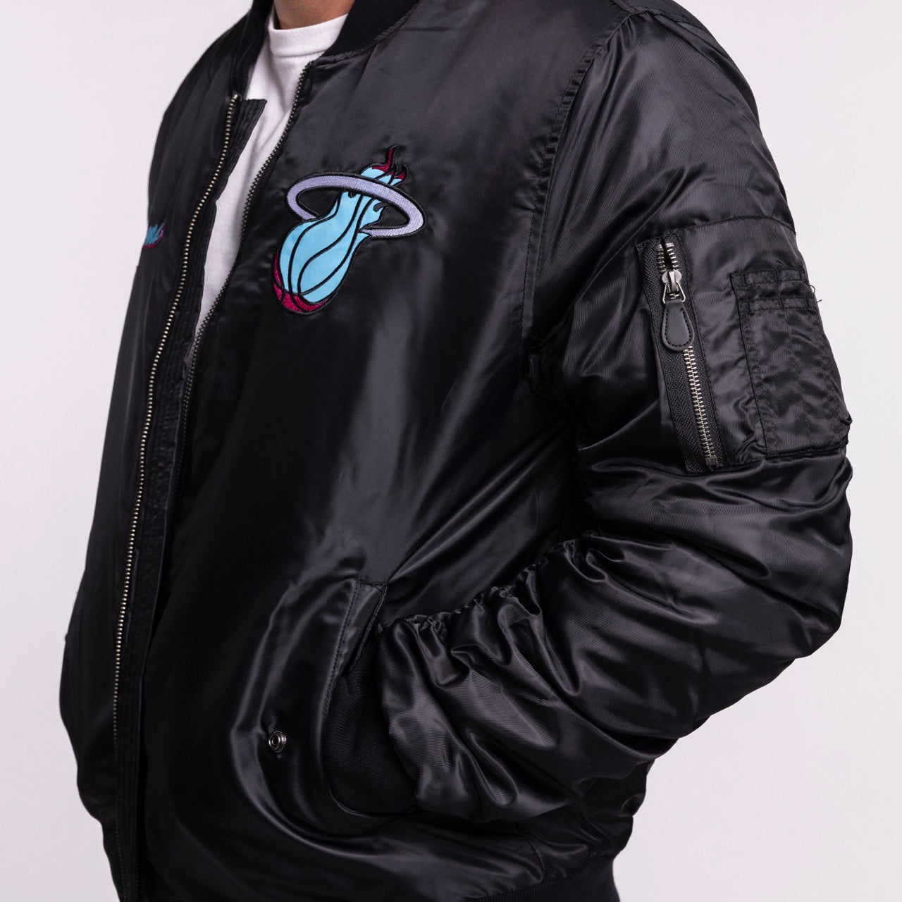 A person wears a men’s size L G-III Sports Miami HEAT Original Vice Nights Aviator Jacket with a shiny black polyester finish, iconic colorful chest logo, visible zipper, sleeve pocket, standard fit, and machine-washable for easy care.