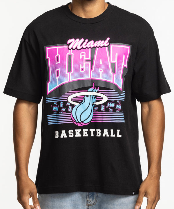 A man wearing a '47 Brand Miami HEAT Original Vice Nights Foundation Tee with colorful retro text and logo stands against a white background in light blue jeans, smiling with one thumb in his pocket. The standard-fit shirt offers all-day comfort.