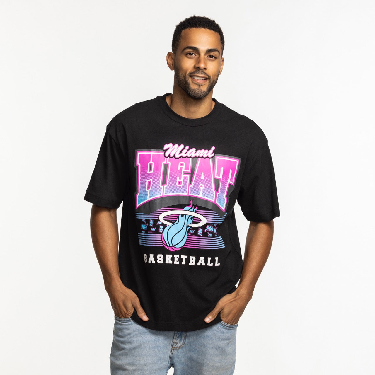A man wearing a '47 Brand Miami HEAT Original Vice Nights Foundation Tee with colorful retro text and logo stands against a white background in light blue jeans, smiling with one thumb in his pocket. The standard-fit shirt offers all-day comfort.