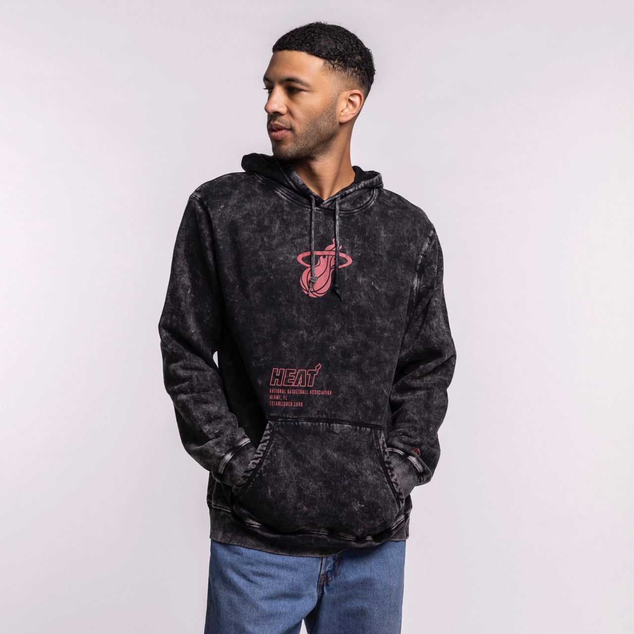 A man wears a New Era Miami HEAT Mineral Wash Logo Hood in black tie-dye with red graphics and blue jeans, standing with hands in the front pocket and looking left against a plain light gray background.