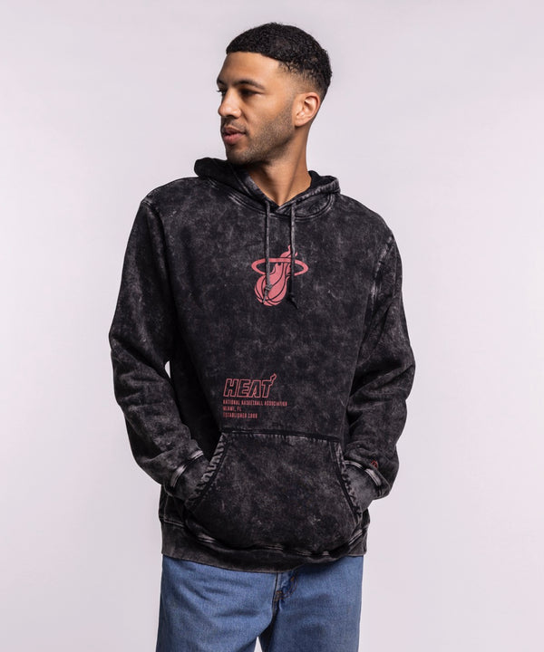 A man wears a New Era Miami HEAT Mineral Wash Logo Hood in black tie-dye with red graphics and blue jeans, standing with hands in the front pocket and looking left against a plain light gray background.