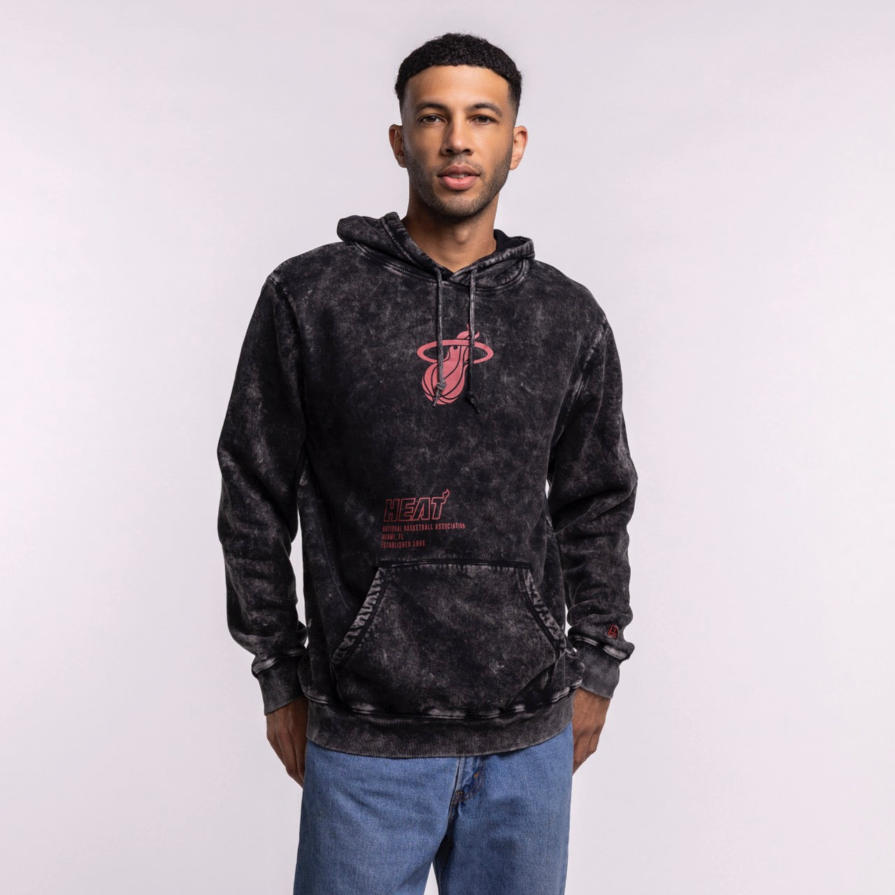 A man stands facing the camera with his hands in his pockets, wearing the New Era Miami HEAT Mineral Wash Logo Hood. This relaxed fit cotton-polyester hoodie features a red team logo and pairs well with light blue jeans against a plain background.