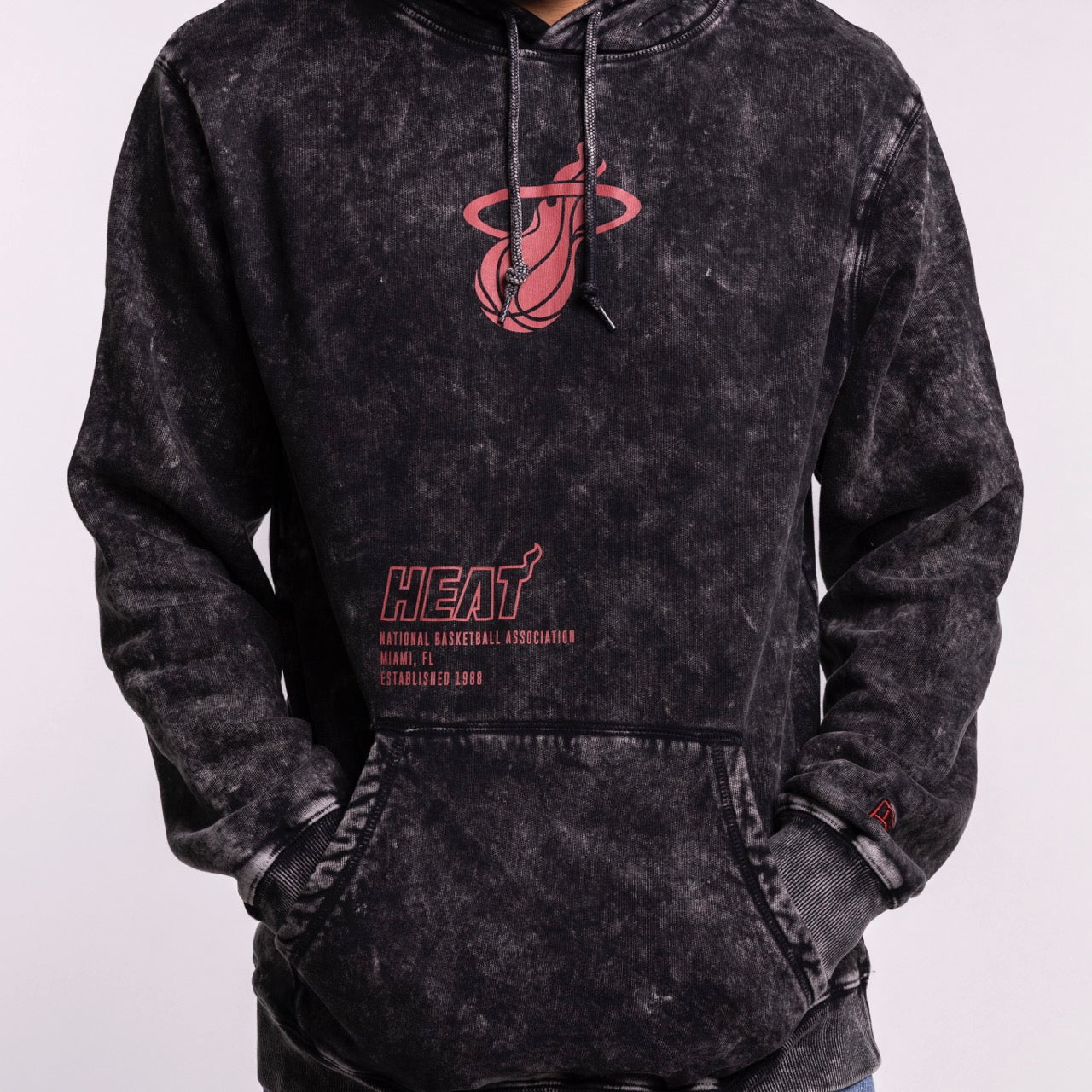 A person wears the New Era Miami HEAT Mineral Wash Logo Hood, a relaxed fit, black washed hoodie with a red Miami Heat logo and text on the front. Their hands are in the pocket. Plain light gray background. Machine wash cold recommended.