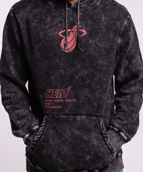 A man wears a New Era Miami HEAT Mineral Wash Logo Hood in black tie-dye with red graphics and blue jeans, standing with hands in the front pocket and looking left against a plain light gray background.
