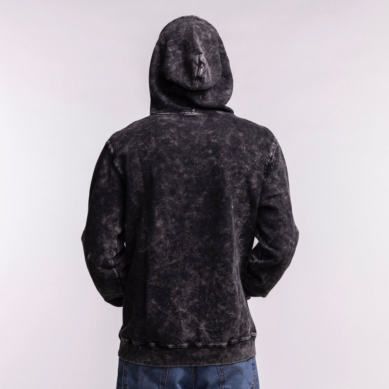 A person wears the New Era Miami HEAT Mineral Wash Logo Hood in washed black, hood up, relaxed fit, with blue jeans, viewed from the back against a plain light gray background. Cotton-polyester with a faded texture. Machine wash cold recommended.
