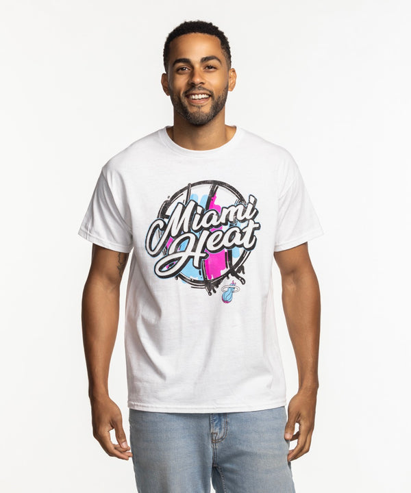 A man smiles against a white background, wearing the Item Of The Game Miami HEAT Original Vice Nights White Tee and light blue jeans.