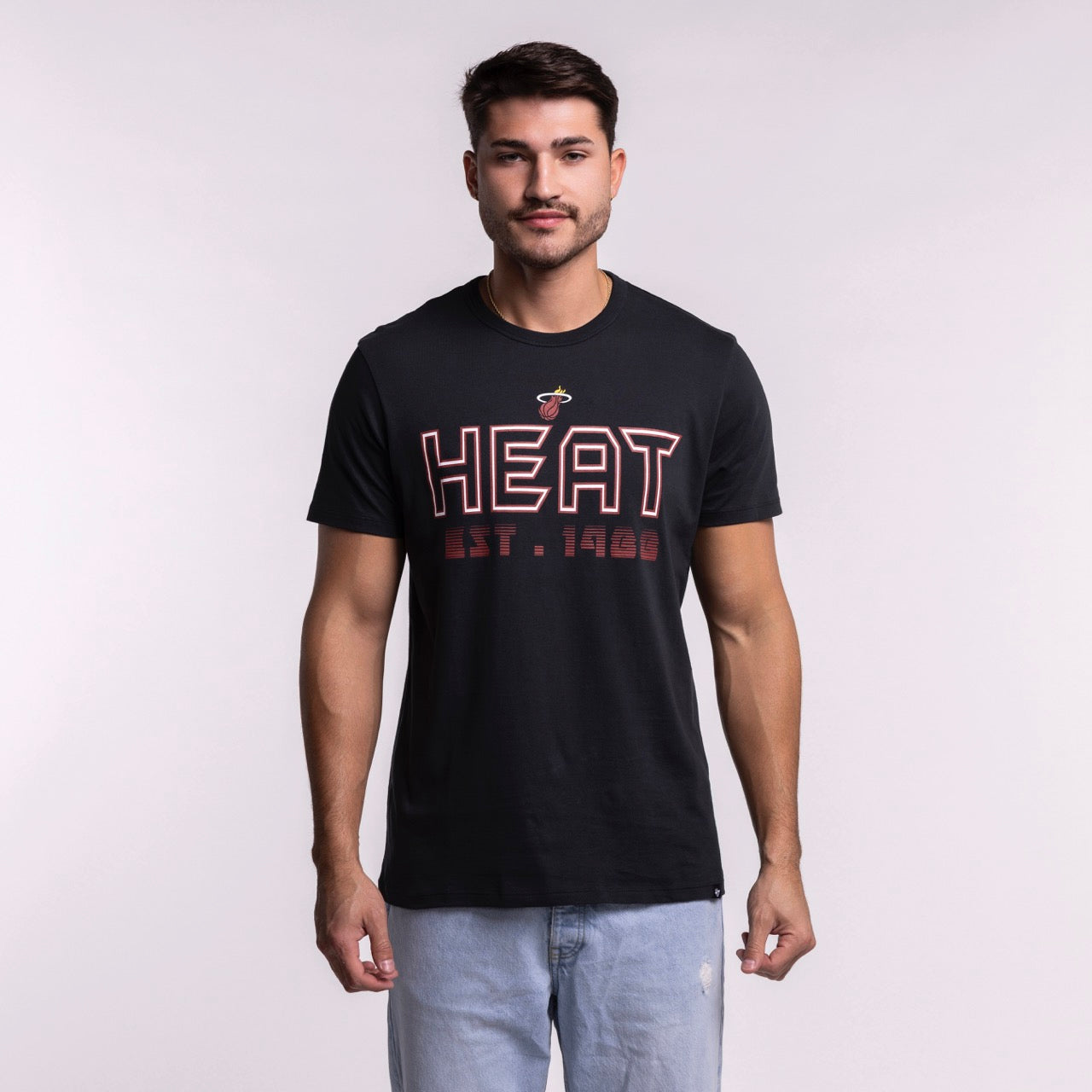 A man wears the '47 Brand Miami HEAT Byline Franklin Tee, a black 100% cotton shirt with HEAT EST. 1988 in red and white, paired with standard fit light blue jeans, against a plain light background. Machine wash cold recommended.