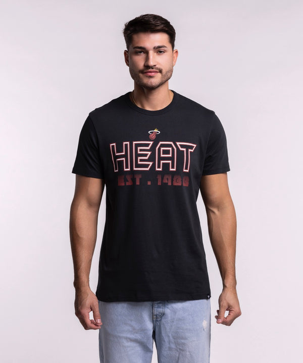 A man wears the '47 Brand Miami HEAT Byline Franklin Tee, a black 100% cotton shirt with HEAT EST. 1988 in red and white, paired with standard fit light blue jeans, against a plain light background. Machine wash cold recommended.