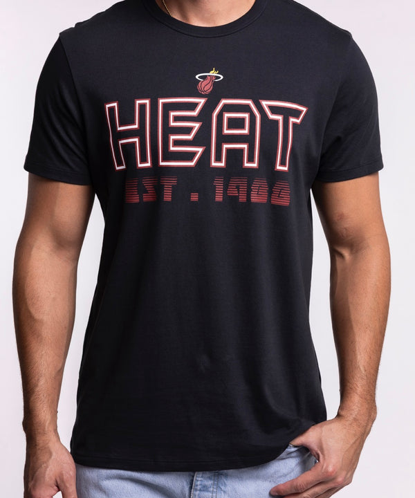 A man wears the '47 Brand Miami HEAT Byline Franklin Tee, a black 100% cotton shirt with HEAT EST. 1988 in red and white, paired with standard fit light blue jeans, against a plain light background. Machine wash cold recommended.