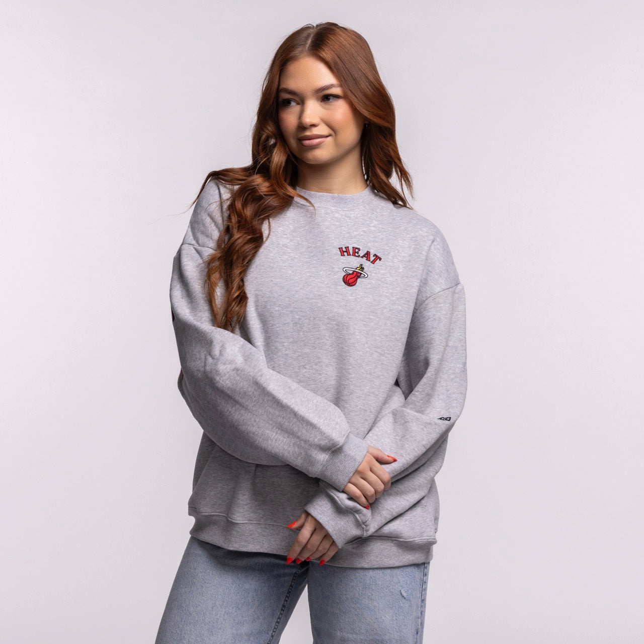 A woman with long brown hair wears the Hype and Vice Miami HEAT Oversized Crew by Hype and Vice, a gray cotton-polyester blend sweatshirt, paired with light blue jeans, standing against a plain background. Hand wash cold recommended.