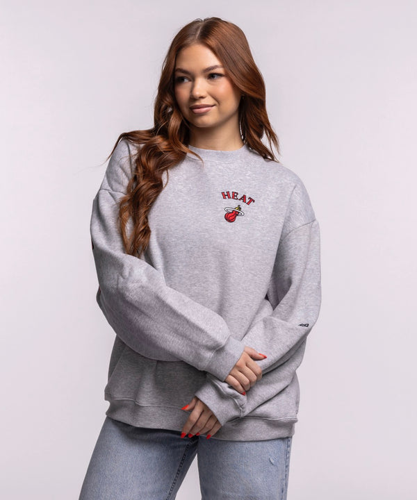 A woman with long brown hair wears the Hype and Vice Miami HEAT Oversized Crew by Hype and Vice, a gray cotton-polyester blend sweatshirt, paired with light blue jeans, standing against a plain background. Hand wash cold recommended.