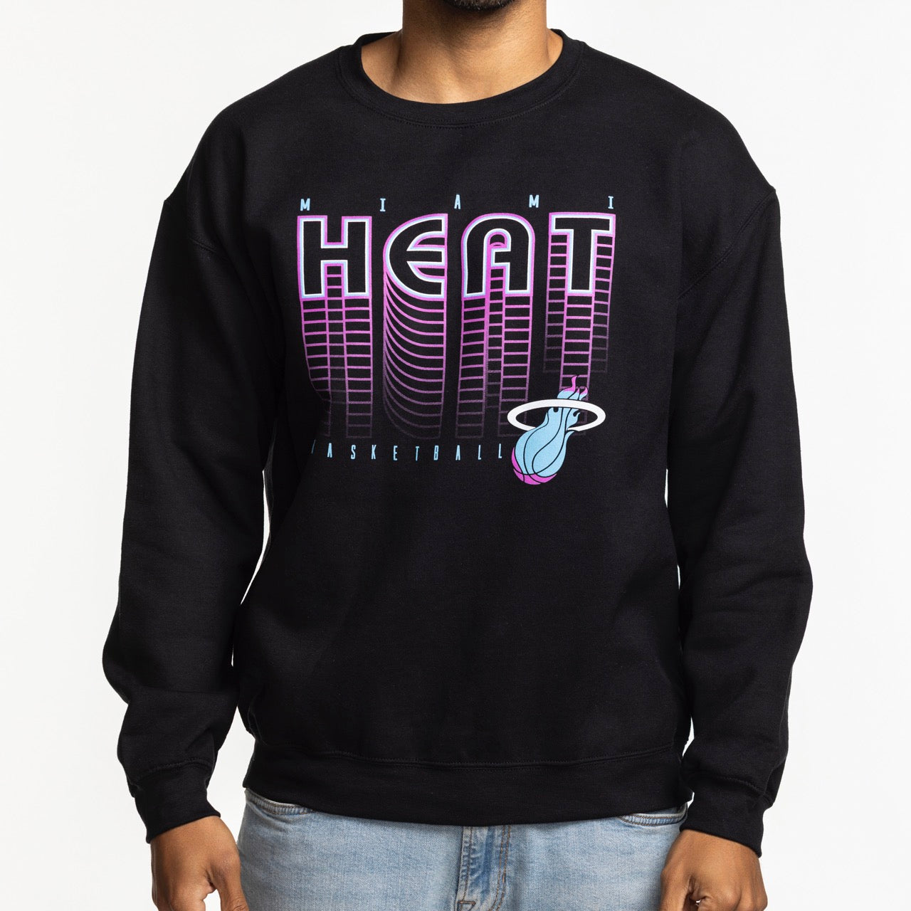 A person wearing the Item Of The Game Miami HEAT Original Vice Nights Retro Crewneck—a standard fit black cotton-polyester sweatshirt with a pink and purple logo and graphic text—stands against a plain white background, torso only visible.
